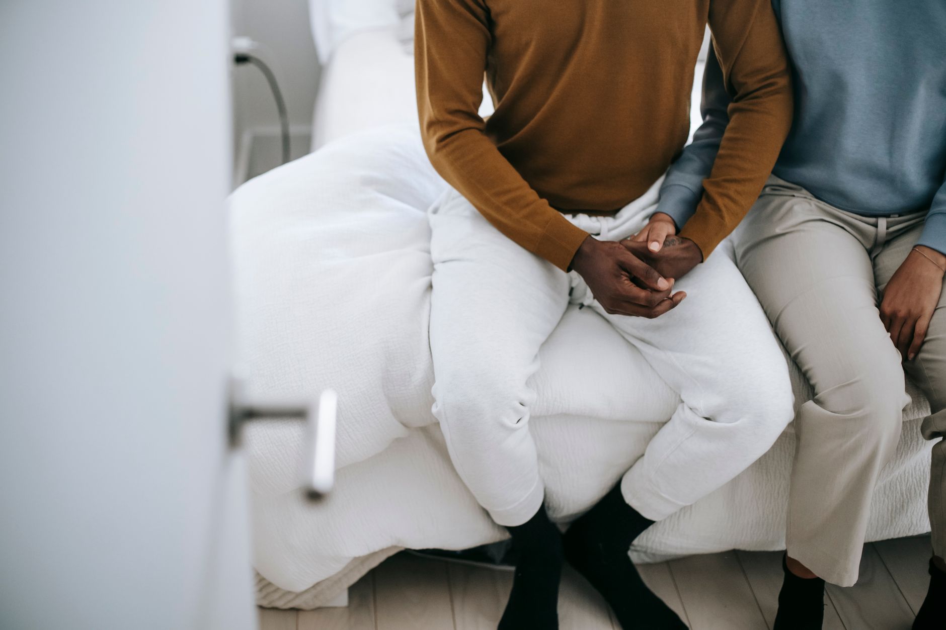 From above of crop anonymous ethnic couple in casual apparel holding hands while sitting in bedroom