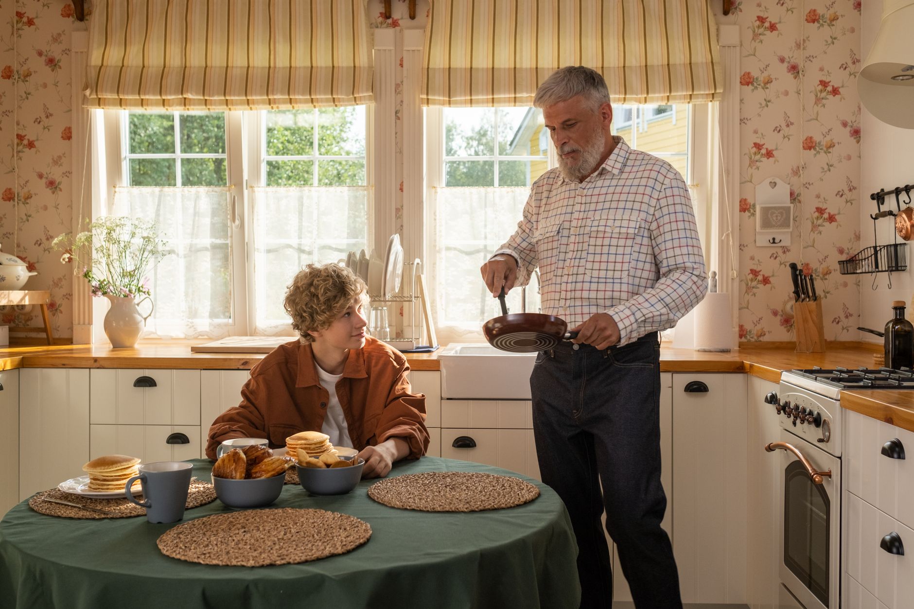 A senior man and teenage boy in a cozy kitchen enjoying pancakes and conversation.