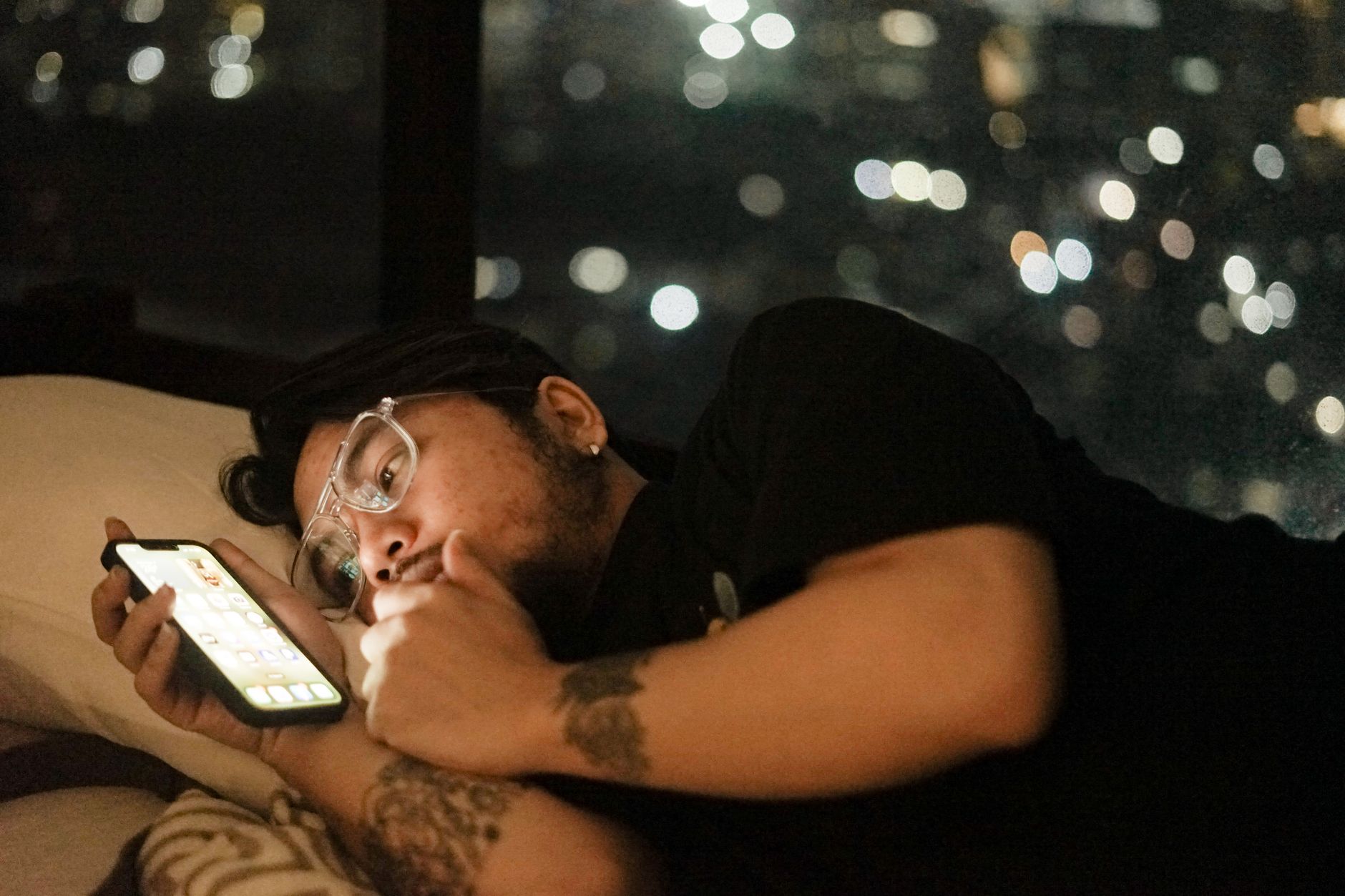 A man with glasses lies in bed viewing his smartphone under dim lighting at night.