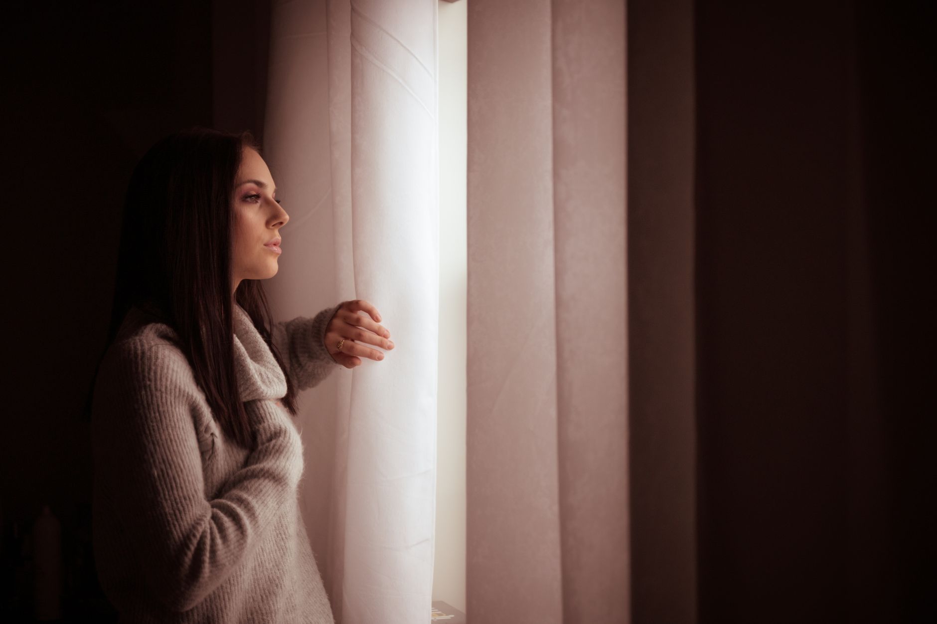 A woman in a sweater thoughtfully gazes out of a sunlit window, creating a warm indoor atmosphere.