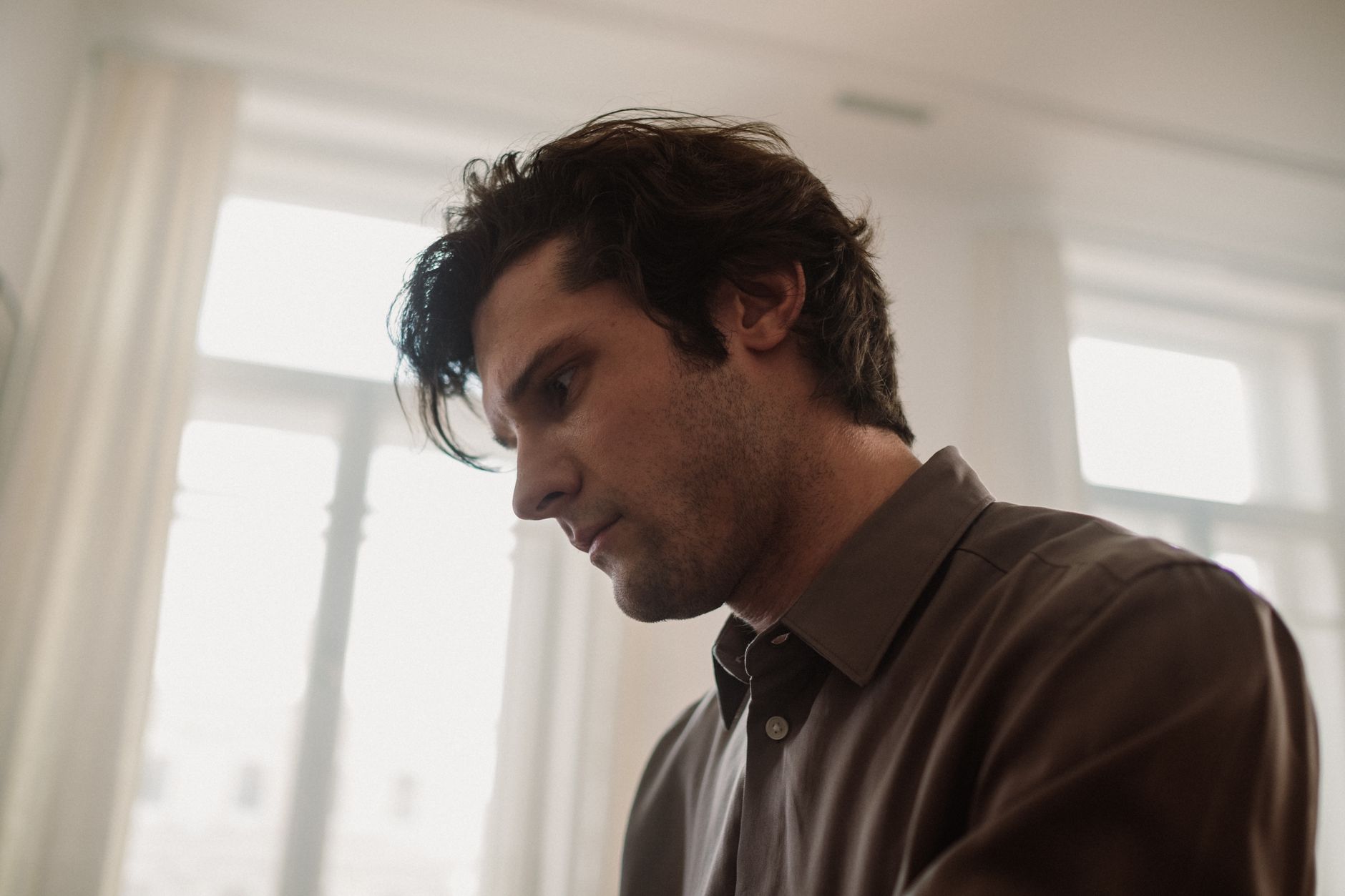 A young man gazing downward in a softly lit room with a thoughtful expression.