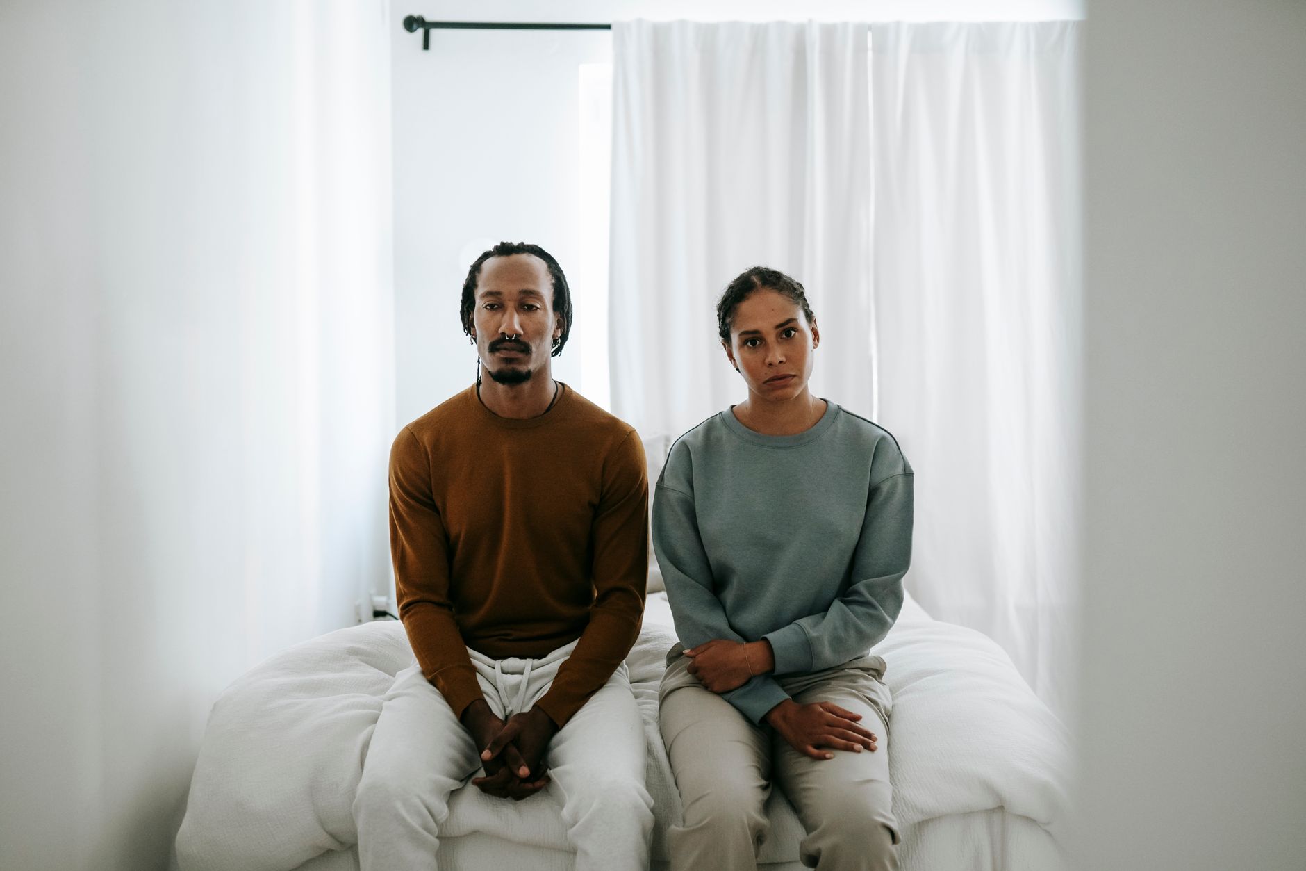 Young displeased African American couple in casual clothes sitting on bed and looking at camera during dispute