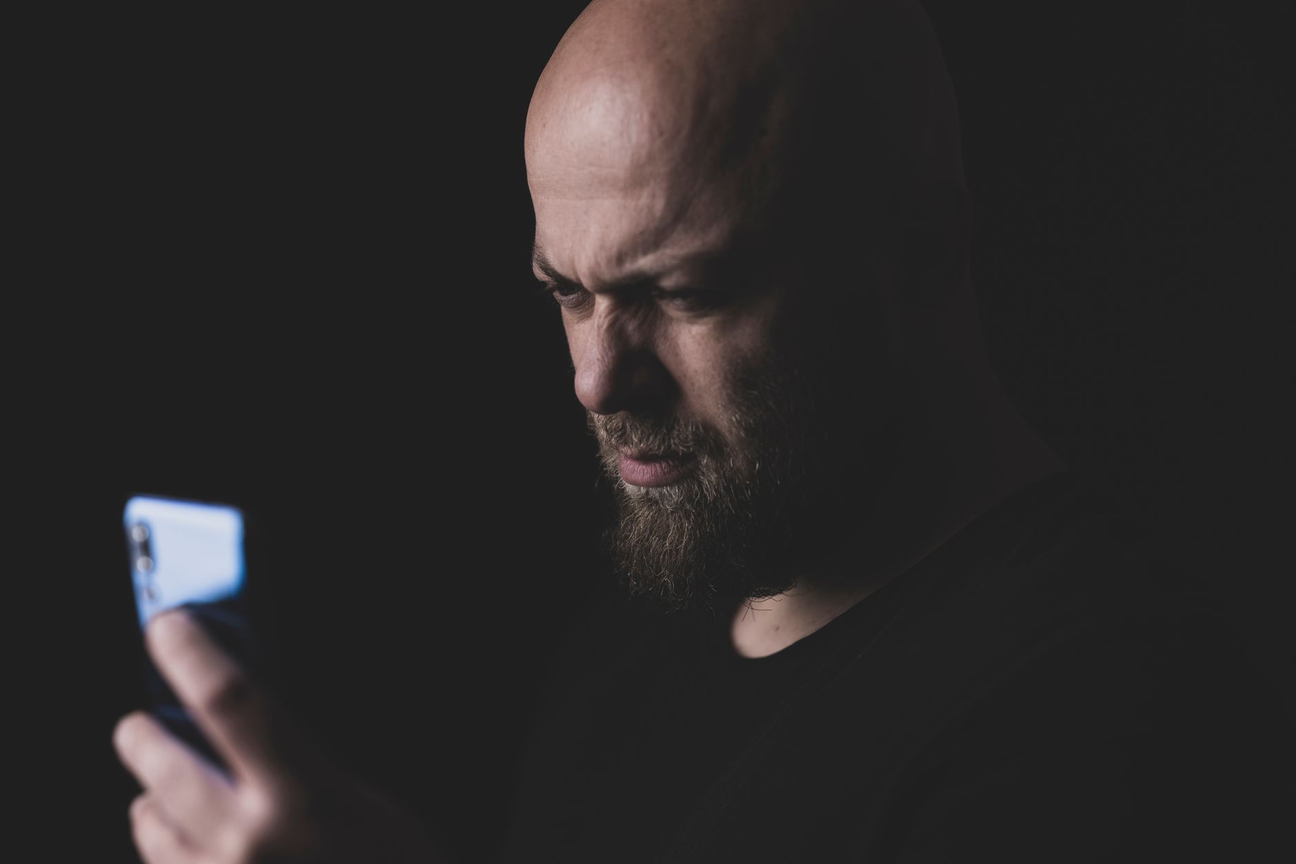 Close-up of a bald, bearded man intently looking at his phone in a dimly lit studio.