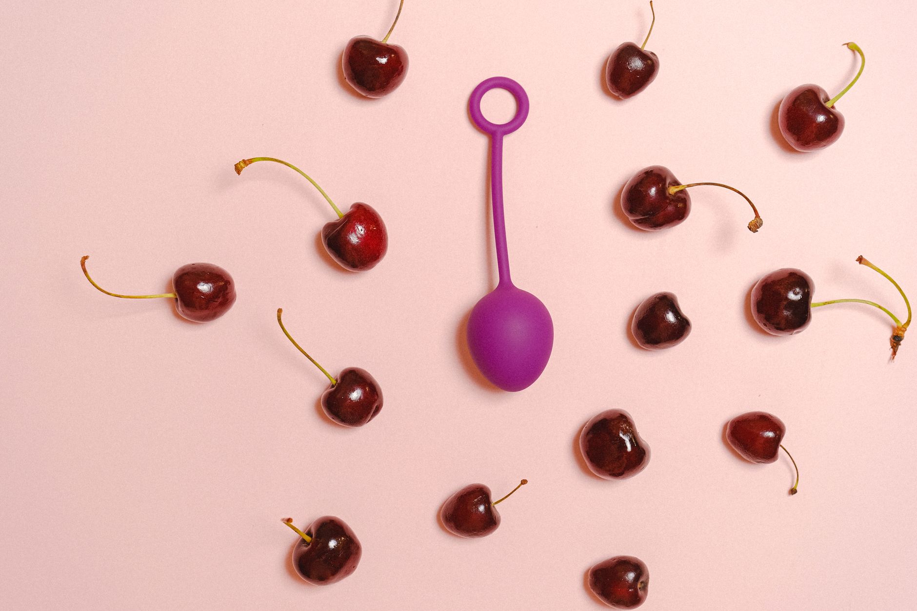 Vibrant still life of a purple silicone toy surrounded by cherries on a pastel pink background.