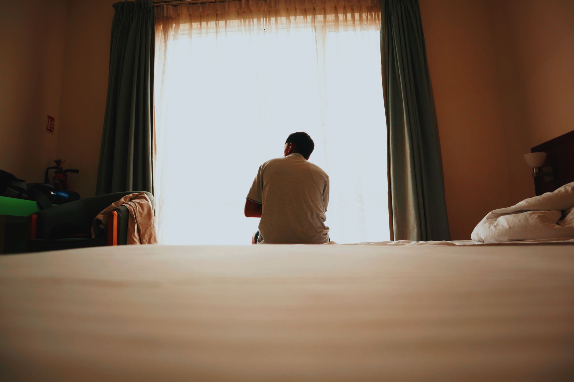 Adult man sitting on bed in cozy bedroom, gazing out bright window.