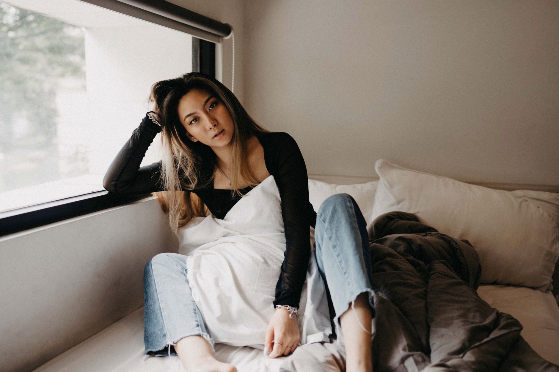 A woman in a black top relaxes by a window in a cozy bedroom, exuding comfort and tranquility.