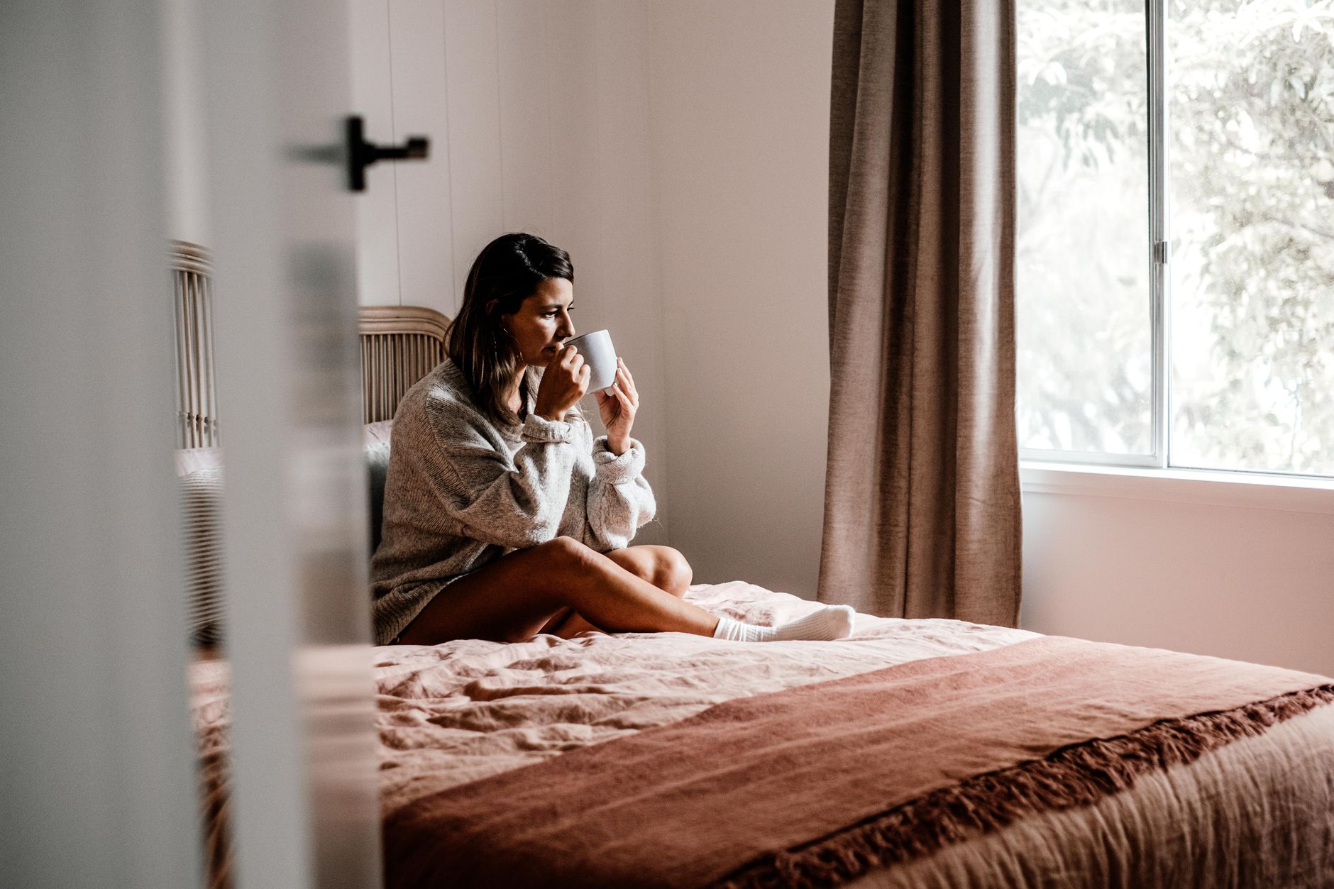 Woman enjoying a hot drink sitting on a bed in a cozy bedroom setting with soft morning light.